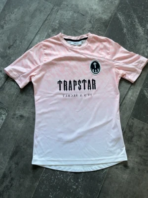 Trapstar t-shirt - Säljer en rosa t-shirt från Trapstar med svart logga och texten 'IT'S A SECRET' på framsidan samt stort '22' och Trapstar-tryck på ryggen. T-shirten har korta ärmar och är gjord i ett mjukt, syntetiskt material med subtilt mönster.