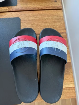 Moncler slides i rött, vitt och blått - Snygga slides från Moncler med bred rem i rött, vitt och blått samt präglad logga framtill. Sulan är svart och har en strukturerad yta för extra grepp. Perfekta för sommaren och enkel att matcha till olika outfits. Köpt på room 1006 på Emporia för 2999 kr mitt pris 1500 kr. Finns i Malmö kan hämtas. Kan också skickas.