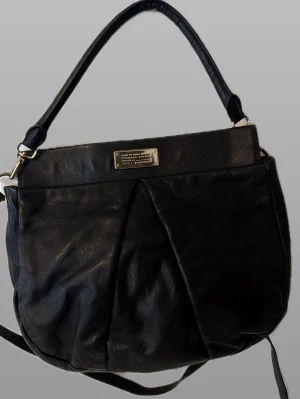 Marc Jacobs väska  - Snygg svart handväska i skinnimitation med dekorativ metallbricka framtill. Väskan har både axelrem och handtag, samt veckad design på framsidan för en trendig look. Perfekt storlek för det viktigaste och passar till många olika outfits.
