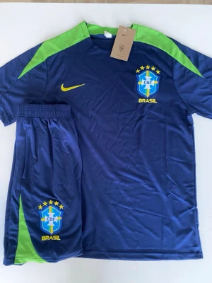 Brasilien fotbollsställ Nike blå/grön - Säljer ett Brasilien fotbollsställ från Nike i blått med gröna detaljer. Setet består av en t-shirt och shorts, båda med CBF-logga och Nike swoosh i gult. Materialet är lätt och andas, perfekt för träning eller match. Stället har elastisk midja på shortsen.