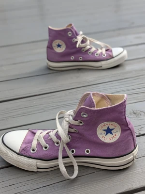 Lila Converse All Star high tops - Säljer ett par lila Converse All Star high tops med klassisk vit tå och sula. Skorna har vita snören, silverfärgade öljetter och den ikoniska loggan på sidan. Tillverkade i canvas med gummisula. Perfekta för dig som vill sticka ut med färg!