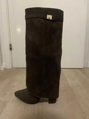 Bruna knähöga mockastövlar - Snygga bruna knähöga stövlar i mocka med spetsig tå. Stövlarna har ett dekorativt spänne upptill i guldton och en stilren, minimalistisk design. Perfekta för att lyfta din outfit under höst och vinter.