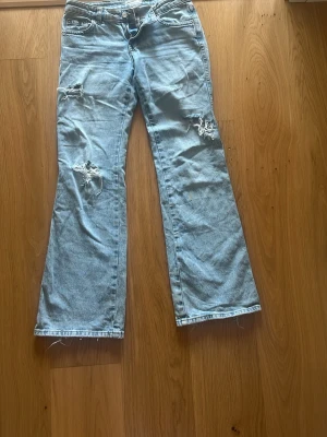 Ljusblå slitna bootcut jeans med hål - Säljer ett par ljusblå jeans med bootcut passform och slitningar på både lår och knän. Jeansen har klassisk femficksdesign och låg midja. 