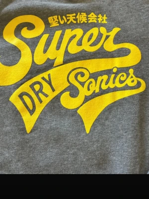 Grå hoodie med gul Superdry-tryck - Snygg grå hoodie från Superdry med stort gult tryck på bröstet där det står 'Super Dry Sonics' och japanska tecken. Hoodien har huva med dragsko, magficka och ribbade muddar. Perfekt för en avslappnad och trendig stil.