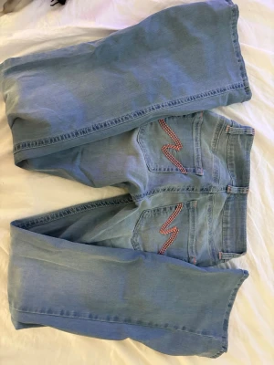 Nelly Jeans - Säljer ett par ljusblå bootcut jeans med snygg rödvit broderad detalj på bakfickorna. Jeansen har klassisk femficksdesign, normal midja och raka ben. Perfekta för dig som gillar retrovibbar och vill sticka ut lite extra. Aldrig använda bara tvättade, därför ingen prislapp kvar.