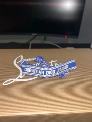 Blått Christian Dior J'adior armband - Snyggt blått tygarmband från Christian Dior med texten 'CHRISTIAN DIOR J'ADIOR' broderad i vitt. Armbandet har justerbar knytning med tofsar i ändarna och en stilren, ungdomlig design som sticker ut. Pris kan diskuteras yterst lite.