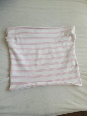 Randig vit och rosa tubtopp - Säljer en söt tubtopp i vitt med ljusrosa ränder. Toppen är axelbandslös och har en bandeauformad passform som sitter tight mot kroppen. Perfekt för varma dagar och enkel att styla med olika outfits.