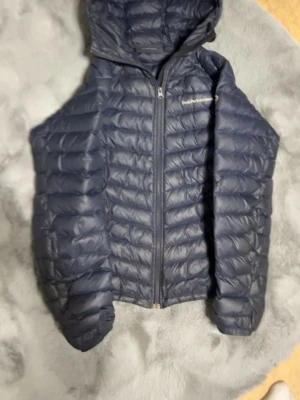 Mörkblå dunväst med huva - Snygg mörkblå dunväst med huva och dragkedja framtill. Västen har quiltad design och är lätt vadderad för extra värme. Perfekt att slänga över en hoodie eller tröja när det är kyligt ute. Materialet är glansigt och ger en modern look.