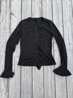Svart ribbad kofta med volangdetaljer - Svart långärmad kofta i ribbat material med knappar framtill och volangdetaljer längs knappraden och vid ärmsluten. Snygg och figurnära passform, perfekt att styla till jeans eller kjol.