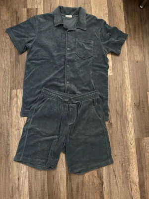 Mörkgrått velourset med skjorta och shorts - Säljer ett mjukt och bekvämt set i mörkgrå velour. Setet består av en kortärmad skjorta med krage, knappstängning och bröstficka samt matchande shorts med resår i midjan och sidofickor. Perfekt för en avslappnad look hemma.