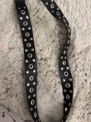 Svart bälte med nitar och hål - Säljer ett svart bälte i syntetmaterial med dubbla rader av silverfärgade nitar och hål längs hela bandet. Perfekt accessoar för att ge din outfit en edgy vibe. Spännet är också i silverfärg och bältet är justerbart.