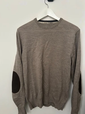Beige stickad tröja med armbågslappar ( stenströms)  - Snygg beige stickad tröja med rund halsringning och mörkbruna armbågslappar. Tröjan har ribbade muddar vid ärmslut och nederkant. Perfekt för dig som gillar en klassisk och stilren look.