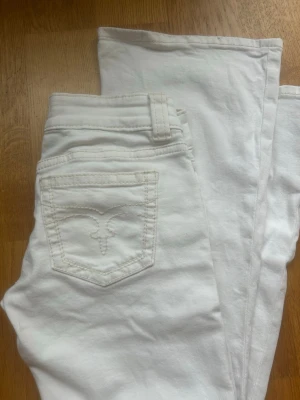 Vita bootcut jeans med broderad ficka - Säljer ett par vita bootcut/ flare jeans med snygg broderad detalj på bakfickan. Jeansen har klassisk femficksmodell, silverfärgade knappar och bälteshällor. Materialet är stretchigt bomullsdenim som sitter tajt och bekvämt. Jeansen har hål på knäna och högre upp som finns inringat på bilden. De är näst in till nya och det finns lite lätt ”uppskrapat” läbgst ner🥰 skruv för fler bilder och frågor😚