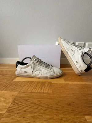 Vita sneakers från Oliver Cabell - Snygga vita sneakers från Oliver Cabell med svart detalj på hälen och grå snörning. Skorna har en låg profil och är tillverkade i läder med en klassisk rund tå och diskret logga på sidan. Perfekta för en clean och stilren look.