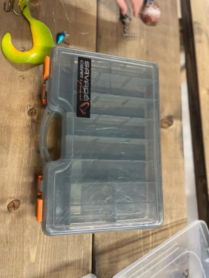 Savage Gear beteslåda med fack - Praktisk genomskinlig beteslåda från Savage Gear med flera justerbara fack och orangea lås. Perfekt för att organisera fiskedrag och småprylar. Tillverkad i tålig plast och har ett smidigt handtag för enkel transport.