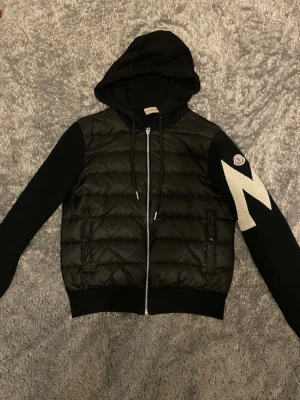 Moncler cardigan!  - Snygg svart pufferjacka med vadderad front, stickade ärmar och stor huva. Dragkedja framtill och två fickor. Ena ärmen har en vit blixtdetalj och en patch. Perfekt för kalla dagar och riktigt trendig look.
