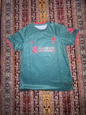 Grön Liverpool matchtröja Nike M - Snygg grön Liverpool matchtröja från Nike med röda detaljer och tryck. Tröjan har korta ärmar, rund hals och är tillverkad i Dri-FIT material som andas. Perfekt för fotbollsträning eller att bära till vardags för fans. Liverpool-logga och sponsortryck på bröstet.