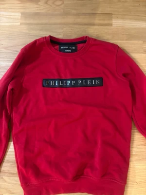 Röd sweatshirt från Philipp Plein - Röd sweatshirt med rund hals från Philipp Plein. Tröjan har en svart patch med logotypen på bröstet och en sexkantig PP-detalj i nacken. Mjuk bomullskänsla och ribbade muddar vid ärmslut och nederkant.