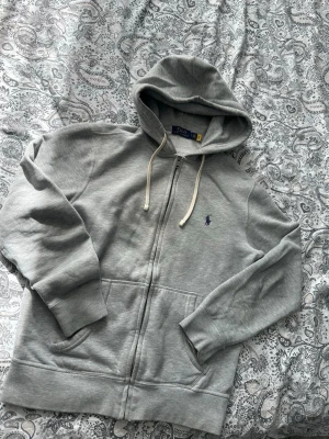 Grå zip hoodie från Polo Ralph Lauren - Snygg grå hoodie med dragkedja från Polo Ralph Lauren. Klassisk design med huva och vita dragsnören, två fickor framtill och den ikoniska lilla blå loggan på bröstet. Perfekt till jeans eller joggers för en chill look.
