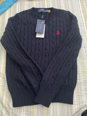 Mörkblå kabelstickad tröja Polo Ralph Lauren - Mörkblå kabelstickad tröja från Polo Ralph Lauren med rund halsringning och röd broderad logga på bröstet. Tröjan har ribbade muddar vid ärmslut och nederkant. Klassisk och tidlös design i mjukt material.