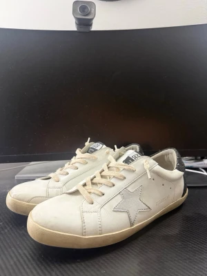 Golden goose  - Snygga vita sneakers med silverfärgad stjärna på sidan och svartglittrig häl. Skorna har beige snören och en klassisk låg profil. Tillverkade i läder med detaljer i mocka och textil. Perfekta för dig som vill ha en trendig och avslappnad look.