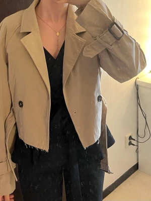 Kort beige trenchcoat med bälte - Säljer en beige cropped trenchcoat med rå kant nertill, klassisk krage och dubbelknäppning framtill. Jackan har ett bälte i midjan och spänne vid ärmsluten för extra detalj. Perfekt för dig som gillar en trendig och avslappnad look.