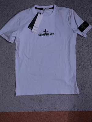 Vit Stone Island t-shirt med logga - Säljer en vit t-shirt från Stone Island med broderad svart logga på bröstet och klassisk patch på ärmen. T-shirten har rund hals och korta ärmar, tillverkad i mjuk bomull. Perfekt för dig som gillar stilrena och trendiga plagg.