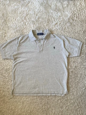 Grå pikétröja från Ralph Lauren - Klassisk ljusgrå pikétröja från Ralph Lauren med korta ärmar och krage. Tröjan har två knappar vid halsen och den ikoniska broderade loggan på bröstet. Perfekt till jeans eller shorts för en clean och stilren look.