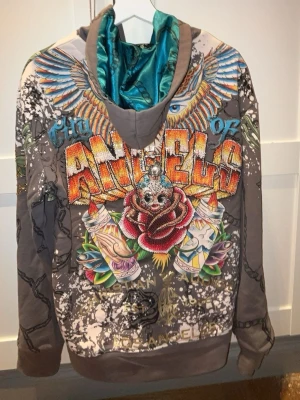 Ed hardy Christian audiger - Unik grå hoodie med stora färgglada tryck på både fram- och baksidan. Framsidan har texten 'City of Angels', en röd ros, vingar och tatuering-inspirerade motiv. Baksidan har gröna dödskallar, rosor och kedjor. Insidan av huvan är turkos. Perfekt för dig som gillar streetstyle och att sticka ut.