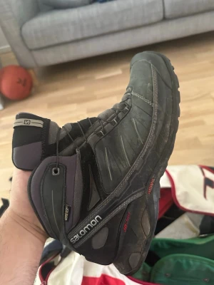 Svarta vandringskängor från Salomon - Säljer ett par svarta och mörkgrå vandringskängor från Salomon med Gore-Tex material. Kängorna har robust sula, hög ankel och snörning för extra stöd. Perfekta för utomhusaktiviteter och äventyr. Salomon-logga på sidan och detaljer i rött.