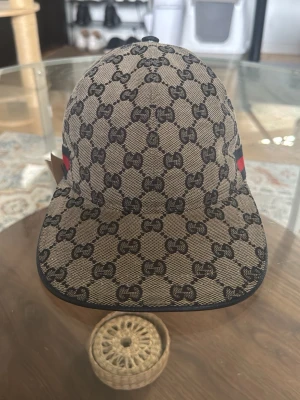 Gucci keps med GG-mönster - Snygg beige keps från Gucci med klassiskt GG-monogram över hela kepsen och svarta detaljer. Baktill finns ett justerbart band och en röd-blå rand. Materialet känns som canvas och kepsen har en böjd skärm. Perfekt för dig som gillar exklusiva accessoarer.