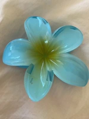 Blå blomformad hårklämma - Säljer en söt hårklämma i form av en blomma med blå och ljusgröna nyanser. Klämman är gjord i plast och har en glansig finish som ger en fräsch och somrig känsla. Perfekt för att piffa upp frisyren med en färgklick!