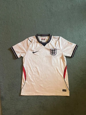 England Nike vit fotbollströja - Säljer en klassisk vit England fotbollströja från Nike med svarta och röda detaljer. Tröjan har korta ärmar, broderad England-logga med stjärna på bröstet och svart Nike-logga. Perfekt för match eller träning.