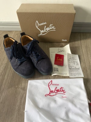 Mörkblå sneakers från Christian Louboutin - Snygga mörkblå sneakers från Christian Louboutin i mocka med klassiska röda sulor. Skorna har blå skosnören, lågt skaft och beige innerfoder. Kommer med originalkartong, dustbag och äkthetsintyg.