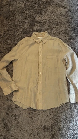 Beige linneskjorta från EAST WEST - Snygg beige skjorta i linne från EAST WEST. Klassisk krage, bröstficka och knäppning framtill. Perfekt för en avslappnad men stilren look. Luftigt material som passar bra till varmare dagar.
