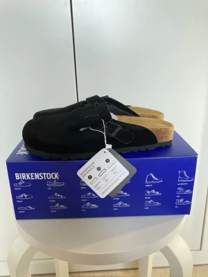 Svarta Birkenstock Boston tofflor - Säljer ett par svarta Birkenstock Boston tofflor i mocka med justerbart spänne och klassisk korksula. Perfekta för dig som vill ha både stil och komfort. Innersulan är mjuk och formad för bästa passform.