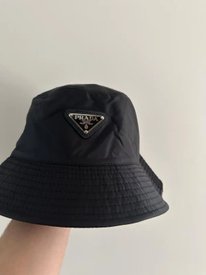 Helt ny svart bucket hat från Prada - Snygg svart bucket hat från Prada med det ikoniska metallmärket framtill. Hatten är tillverkad i ett lätt och slitstarkt nylonmaterial som ger en modern och clean look. Perfekt accessoar för dig som vill addera lite lyx till din outfit. Priset kan diskuteras vid snabb köp!