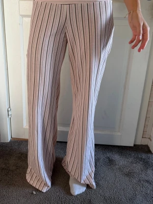 Vida randiga leggings i pastell - Säljer ett par vida leggings med vertikala ränder i ljusrosa, vitt och svart. Mjukt och stretchigt material som sitter skönt i midjan. Perfekta för dig som gillar en avslappnad men trendig look.