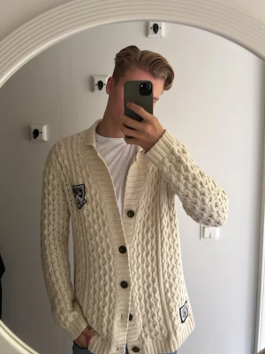 Beige stickad kofta från Bondelid - Säljer en beige stickad kofta från Bondelid i storlek medium. Koftan har snyggt kabelstickat mönster, ribbade muddar och knäppning med stora knappar framtill. På bröstet sitter en Bondelid-patch som ger en preppy känsla. Perfekt för lager-på-lager under kyliga dagar.