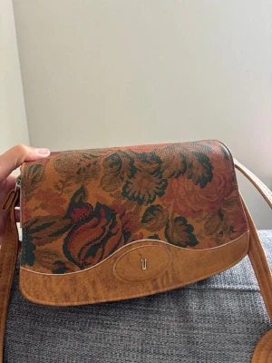 Retro axelväska med blommönster - Säljer en unik axelväska i retrostil med brunt konstläder och färgglatt blommönster i grönt, rött och orange. Väskan har ett lock med metallspänne och justerbar axelrem. Perfekt för dig som gillar vintage och vill sticka ut med din stil. Mått 25x16x10 cm 
