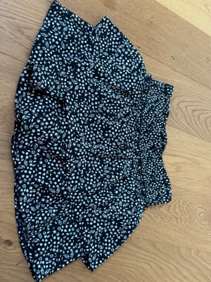 Svart blommig kortkjol med volang - Supersöt svart kortkjol med vitt blommönster och volang. Kjolen har en smockad midja som ger en bekväm och snygg passform. Perfekt att styla med en enkel topp för en trendig look.passar även som xs.