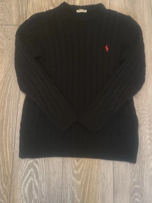 Svart kabelstickad tröja Polo Ralph Lauren - Kabelstickad svart tröja från Polo Ralph Lauren med rund halsringning och röd broderad logga på bröstet. Långa ärmar och ribbade muddar vid ärmslut och nederkant. Perfekt för en stilren och klassisk look.