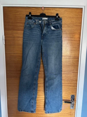Blå raka jeans med hög midja - Säljer ett par klassiska blå jeans med hög midja och raka ben. Jeansen har fem fickor, knappgylf och är gjorda i ett robust denimtyg. Perfekta för en avslappnad och trendig look.