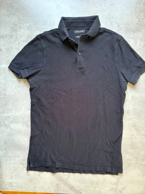 Svart pikétröja från Massimo Dutti - Snygg svart pikétröja från Massimo Dutti med klassisk krage och korta ärmar. Tröjan har diskret struktur i tyget och två knappar vid halsen. Perfekt till en avslappnad men stilren look.