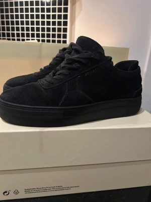 Svarta sneakers i mocka Axel Arigato - Stilrena helsvarta sneakers från Axel Arigato i mjuk mocka. Klassisk modell med tjock sula och svarta skosnören. Perfekta för dig som gillar minimalistisk och clean stil. Diskret logga på sidan och snygg sömnad. Strl 39 i fint skick. Skolåda och påse finns kvar. 