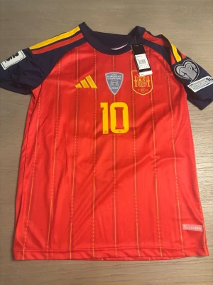 Spansk landslagströja nr 10 röd - Röd fotbollströja från Spaniens landslag med gula detaljer och nummer 10 på både fram- och baksidan. Namnet Lamine Yamal står på ryggen. Tröjan har officiella märken och patchar på ärmarna samt tunna vertikala ränder. Tillverkad i ett lätt funktionsmaterial.