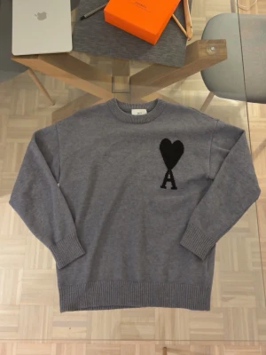 Grå stickad tröja från Ami Paris - Snygg grå stickad tröja från Ami Paris med svart hjärta och bokstaven A broderat på bröstet. Tröjan har rund halsringning, ribbade muddar och är tillverkad i mjukt material. Perfekt för en stilren och avslappnad look. Äkta, kan leta efter kvitton om man frågar.