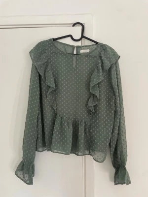 Grön prickig blus med volanger - Söt grön blus i transparent material med små vita prickar och volangdetaljer längs bröstet och axlarna. Blusen har långa ärmar med resår vid handlederna och en lätt utsvängd nederkant.