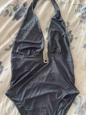 Glittrig mörkgrå body med halterneck - Mörkgrå body i glittrigt material med djup urringning och halterneck. Framtill finns en silverfärgad metall-detalj som ger extra stil. Tyget är stretchigt och följsamt, perfekt för en utekväll på krog eller klubb.