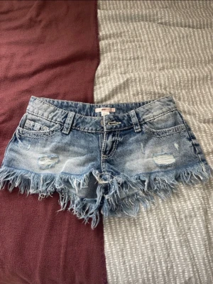 Blå jeansshorts från Gina Tricot - Säljer ett par blå jeansshorts från Gina Tricot i storlek XXS. De har slitna detaljer, fransig kant och klassisk femficksmodell. Perfekta för sommaren och ger en avslappnad vibe till din outfit.